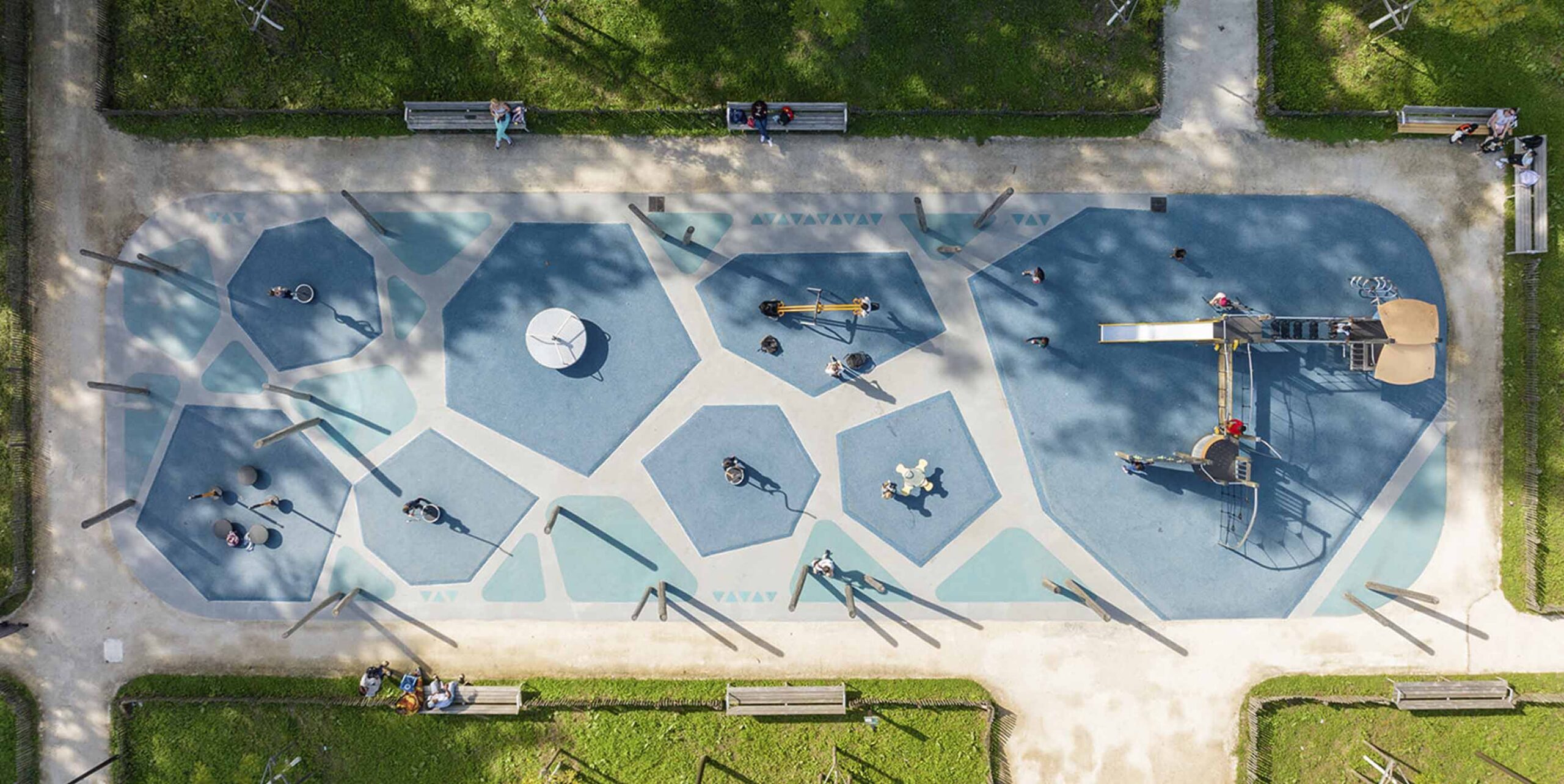 Vue aérienne d'une aire de jeux aménagée au sol bleu géométrique, entourée de pelouse et de bancs.