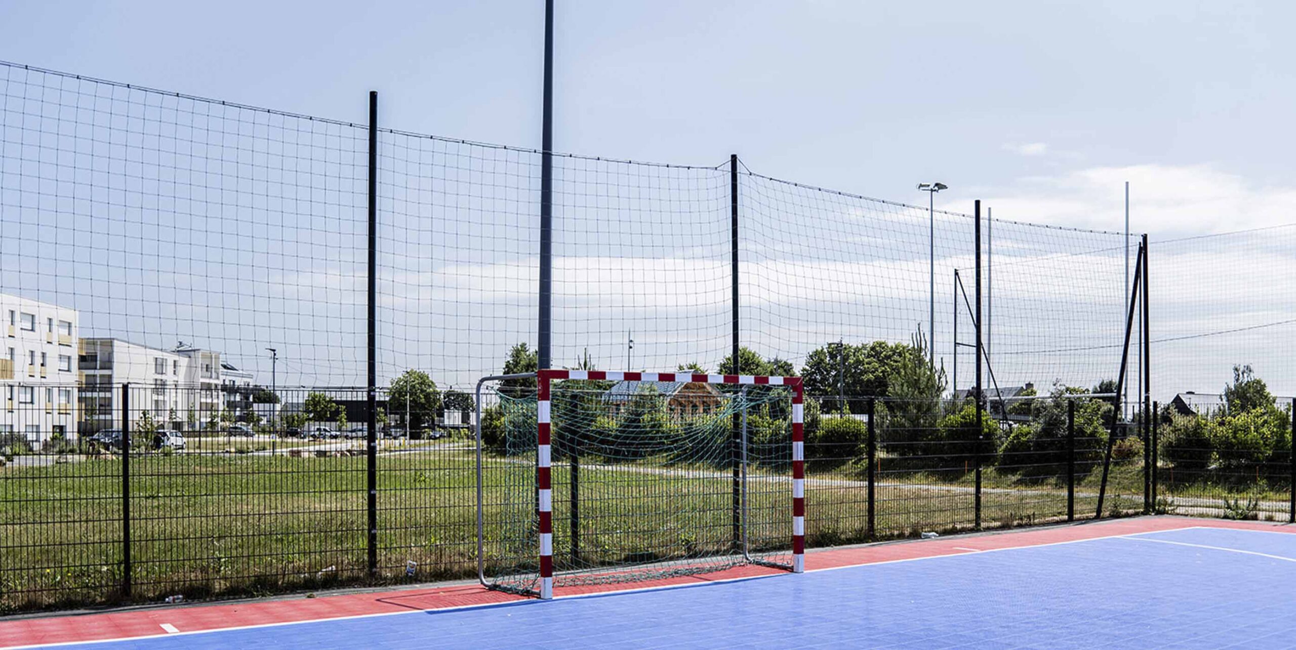 Terrain multisport en résine bleue et rouge entouré d'un espace vert engazonné en zone résidentielle.