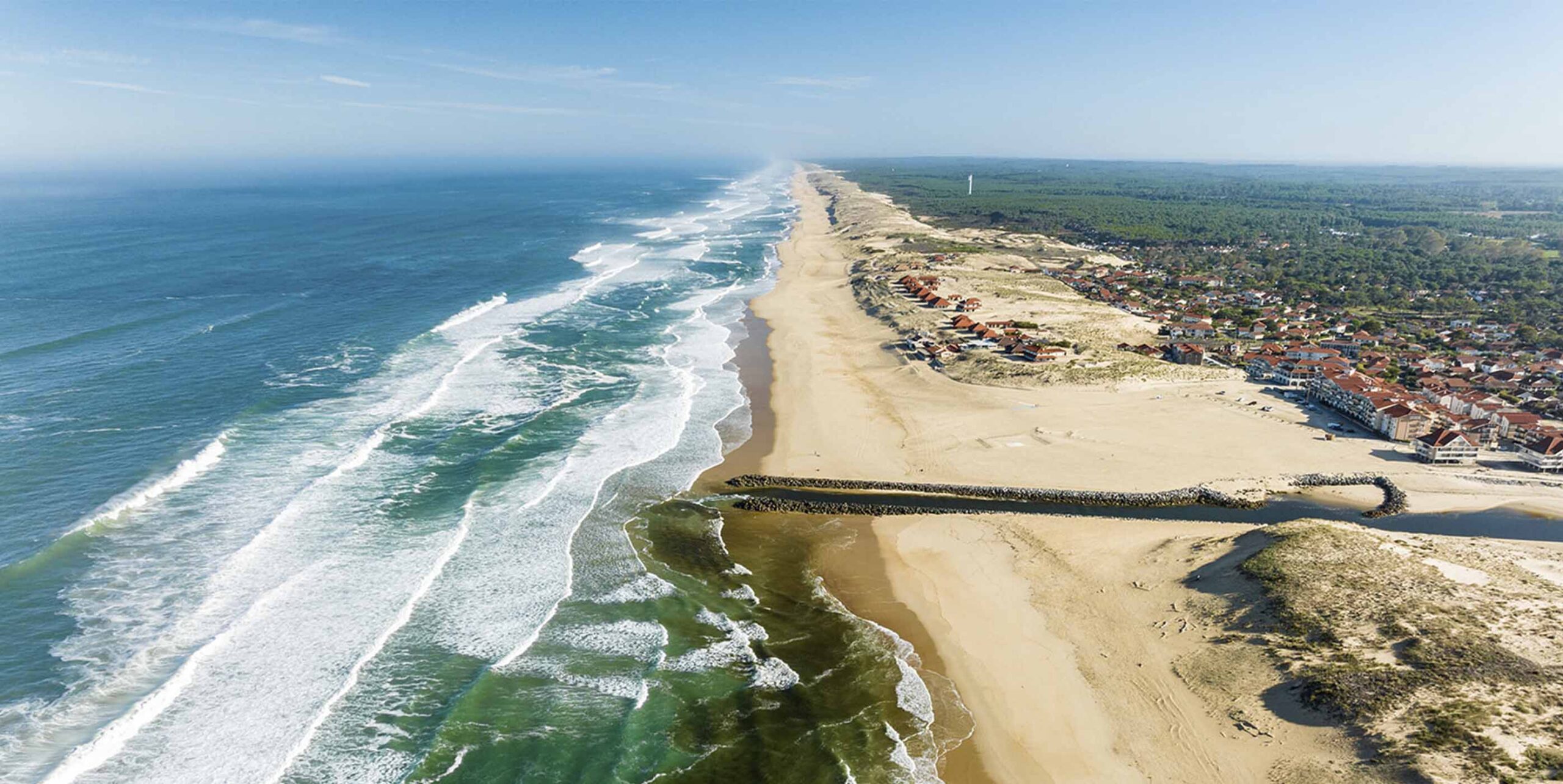 Vue aérienne du littoral atlantique avec plage, dunes et forêt de pins en arrière-plan d'une station balnéaire.