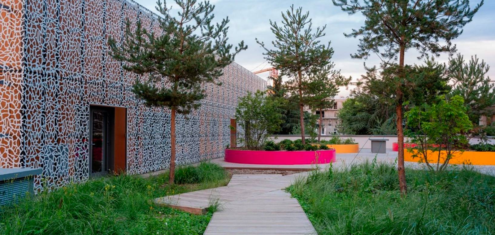 Toiture végétalisée avec pins, prairie fleurie et bacs plantés colorés le long d'une façade urbaine ajourée.