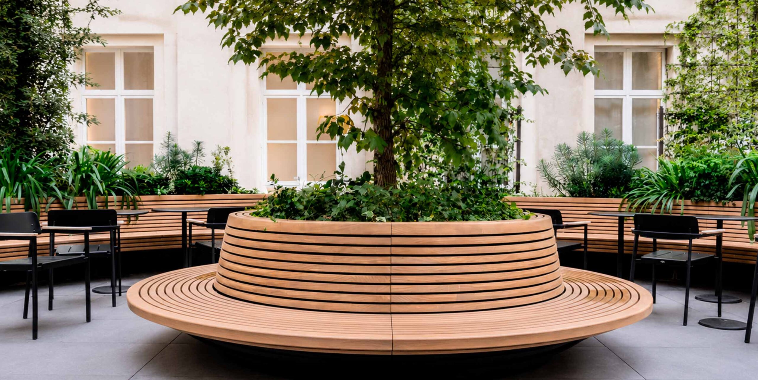 Cour intérieure aménagée avec banquette circulaire en bois entourant un arbre et mobilier de terrasse.