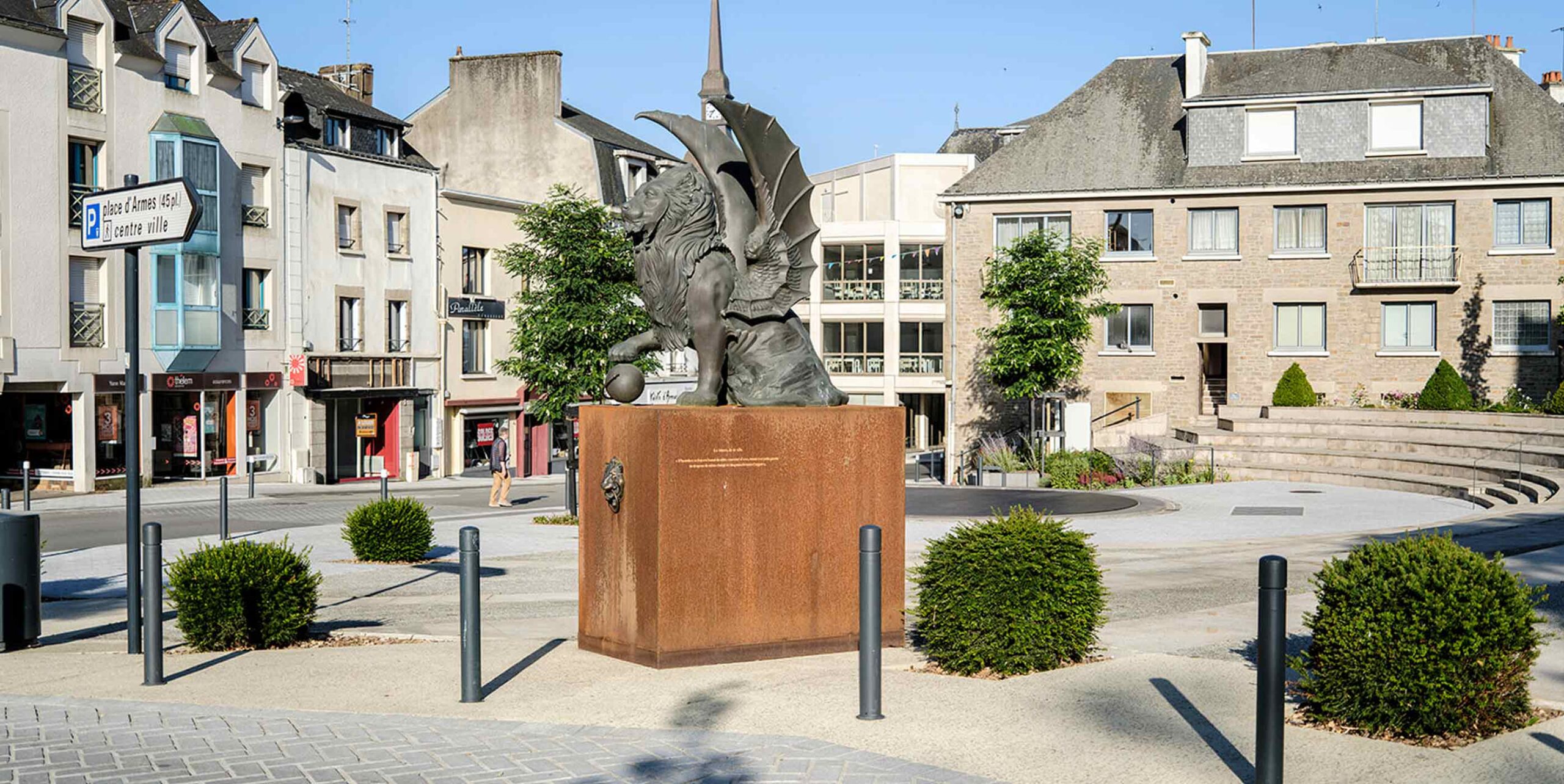 Place urbaine aménagée avec massifs arbustifs taillés en boules autour d'une statue sur socle.