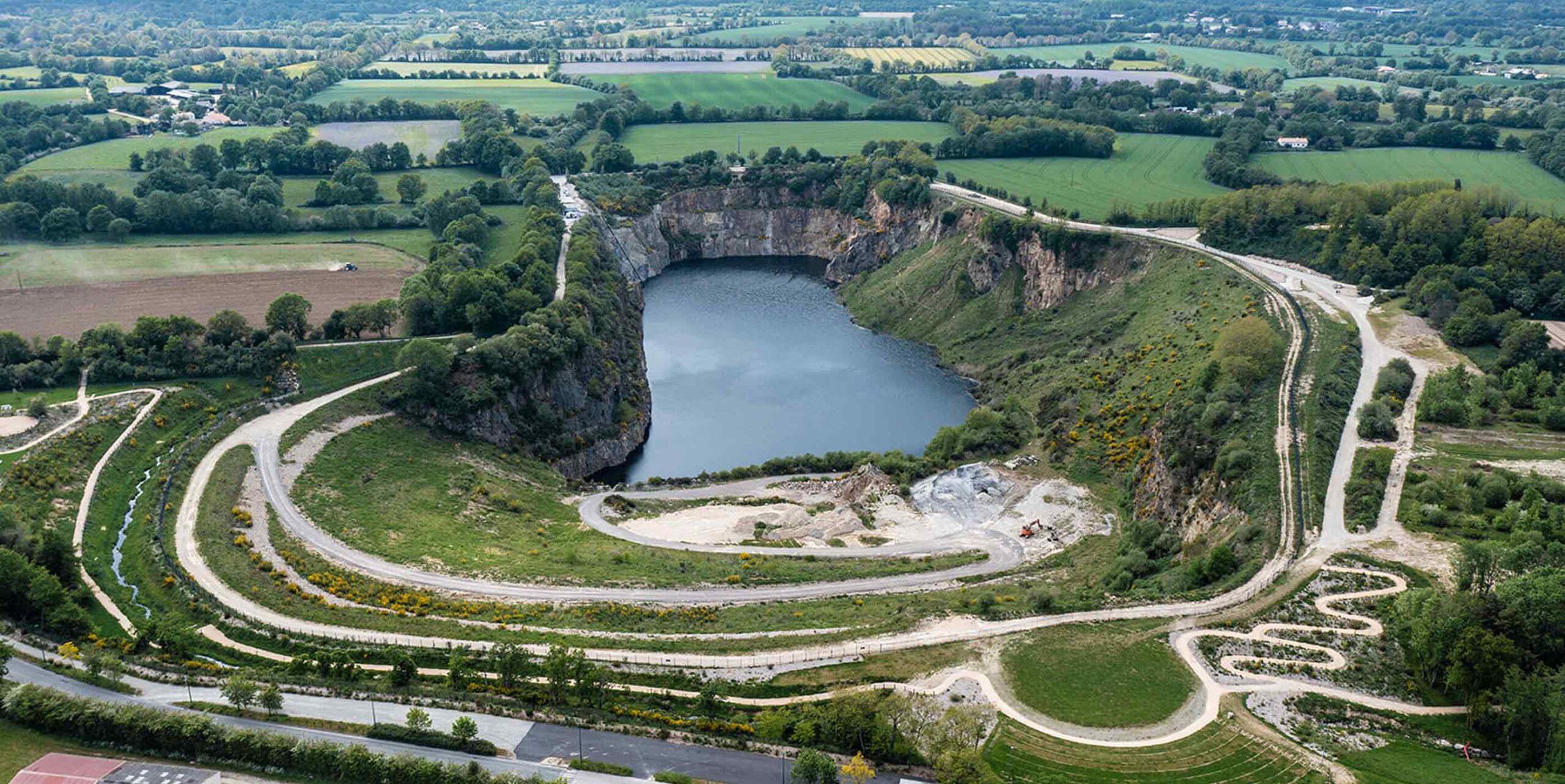Vue aérienne d'une ancienne carrière reconvertie en plan d'eau entourée de chemins aménagés et de végétation.