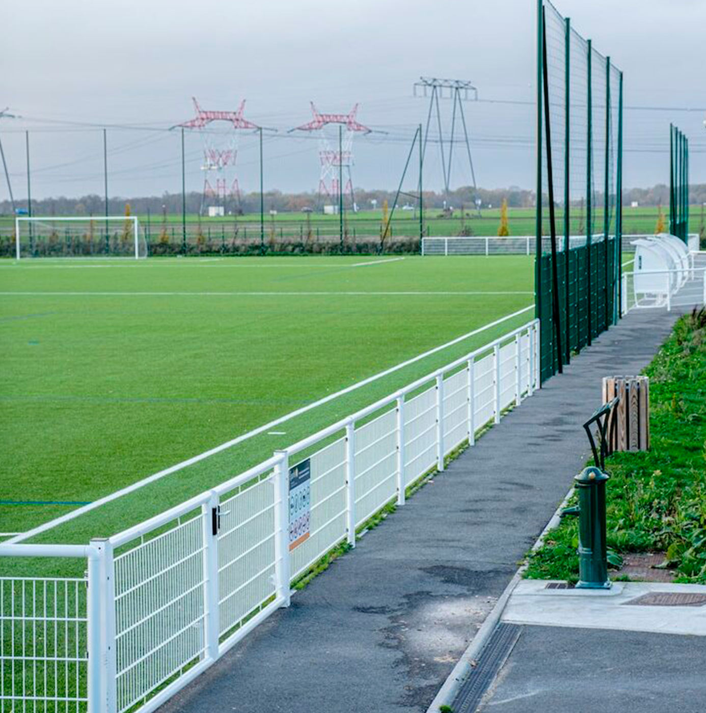 Terrain de football en gazon synthétique clôturé, bordé d'une allée piétonne.