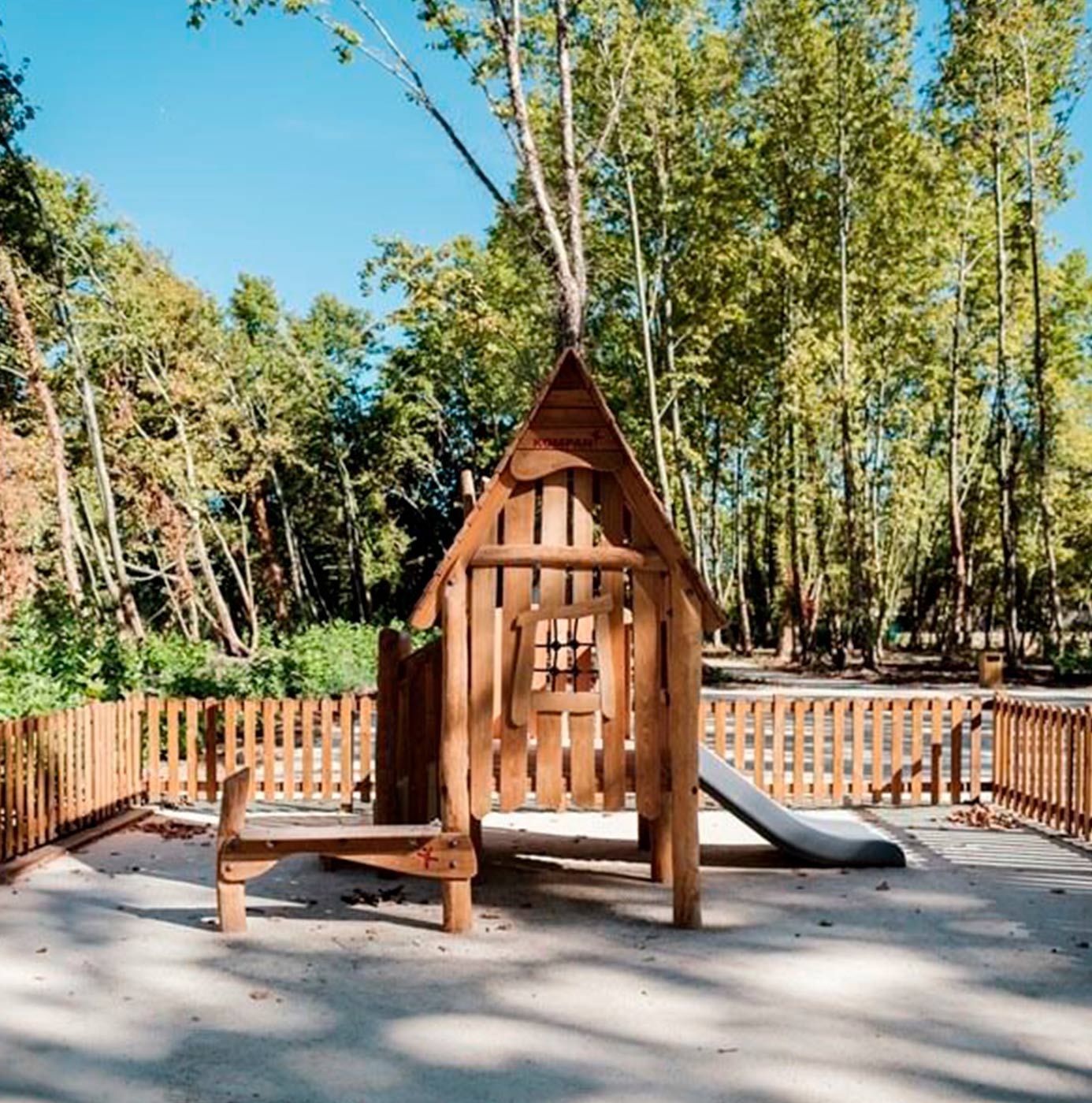 Aire de jeux en bois avec toboggan, clôturée et installée dans un espace boisé.