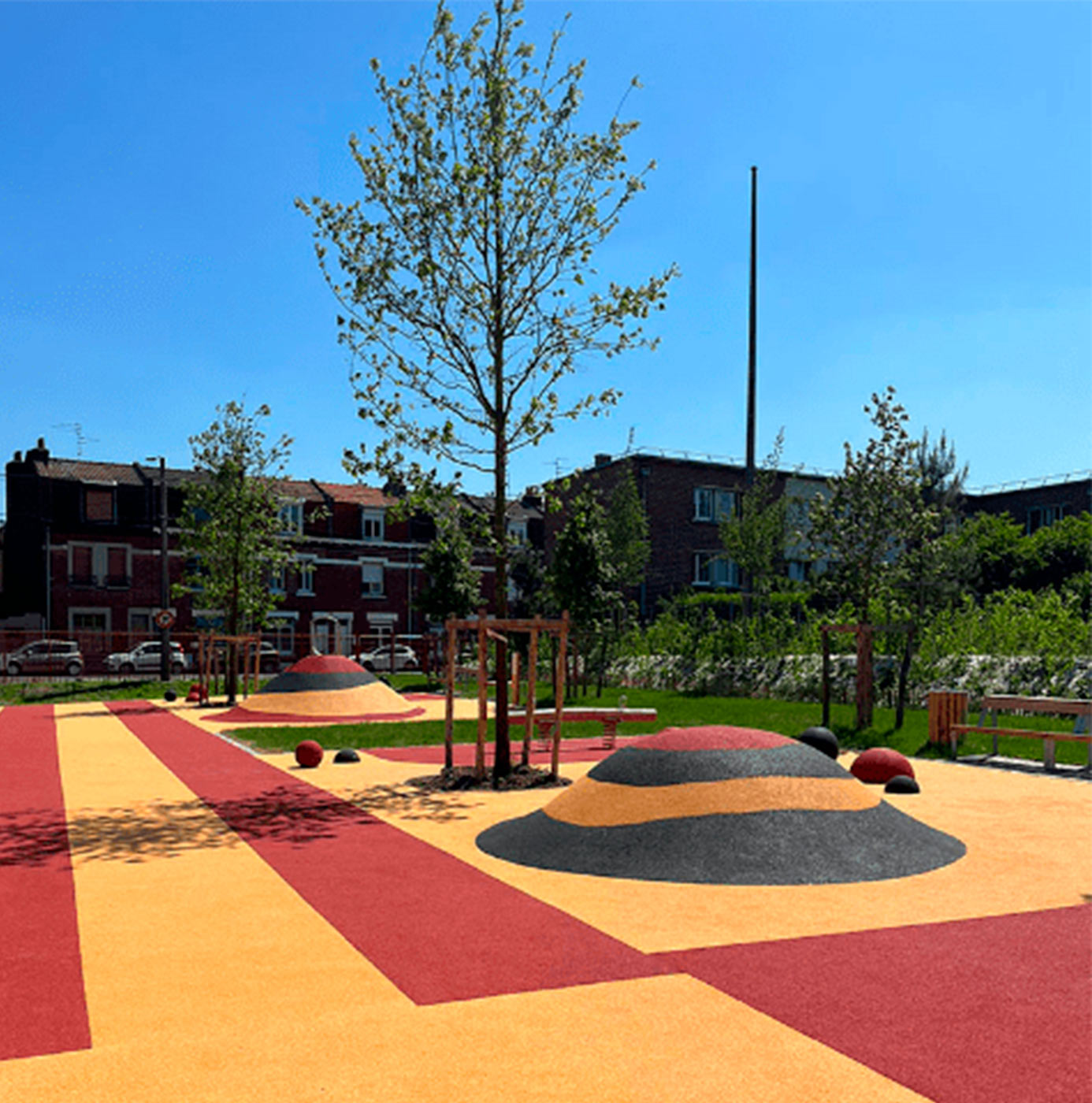 Aire de jeux urbaine avec sol souple coloré, buttes en relief et arbres nouvellement plantés en milieu résidentiel.