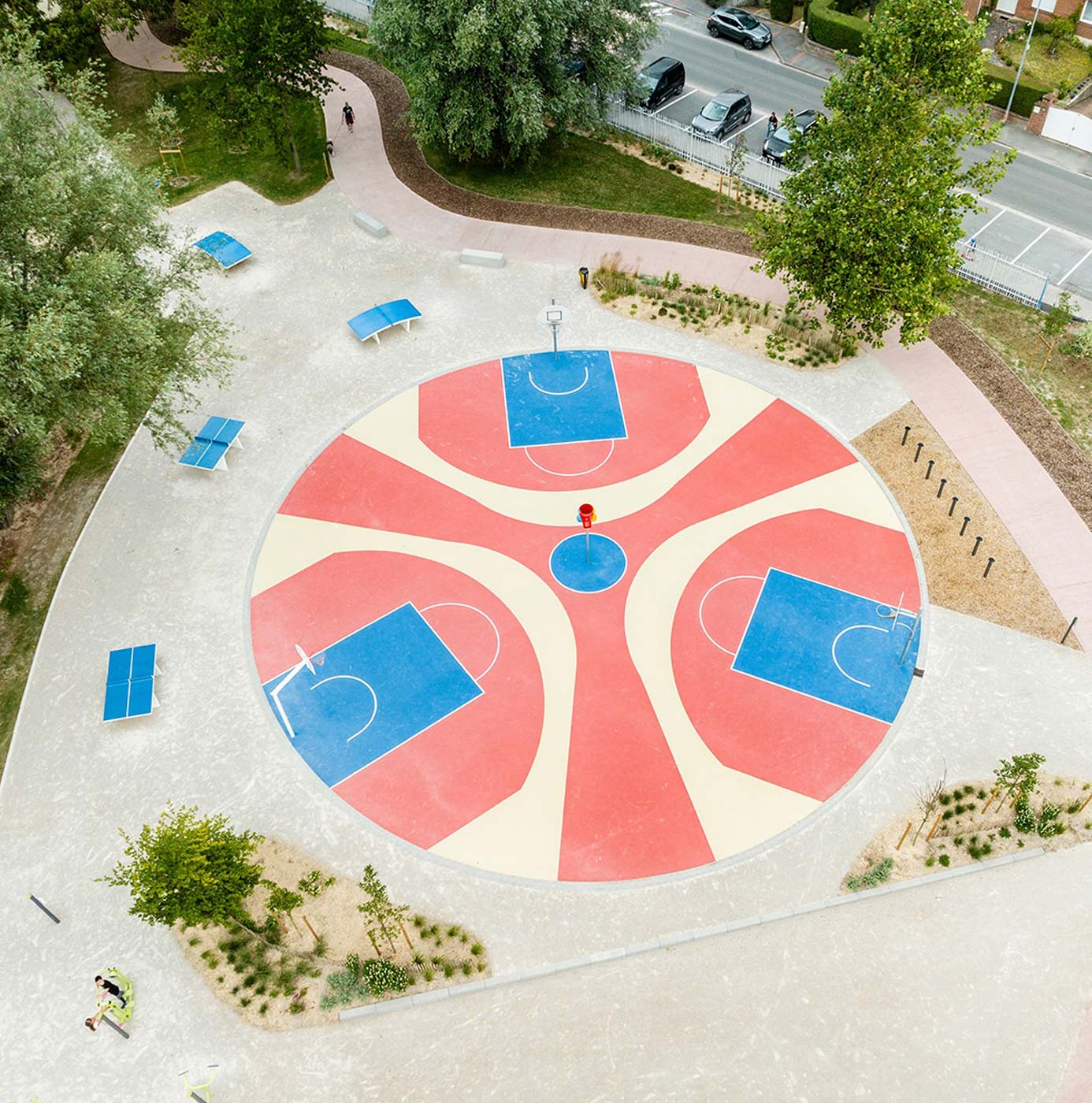 Vue aérienne d'un city stade circulaire à trois terrains de basket entouré d'un parc paysager.