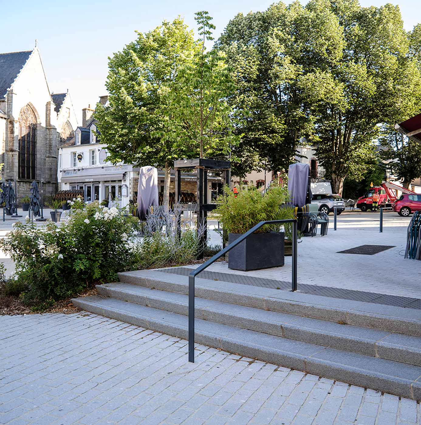 Place urbaine paysagée avec arbres, massifs arbustifs et escalier en granit devant une église.