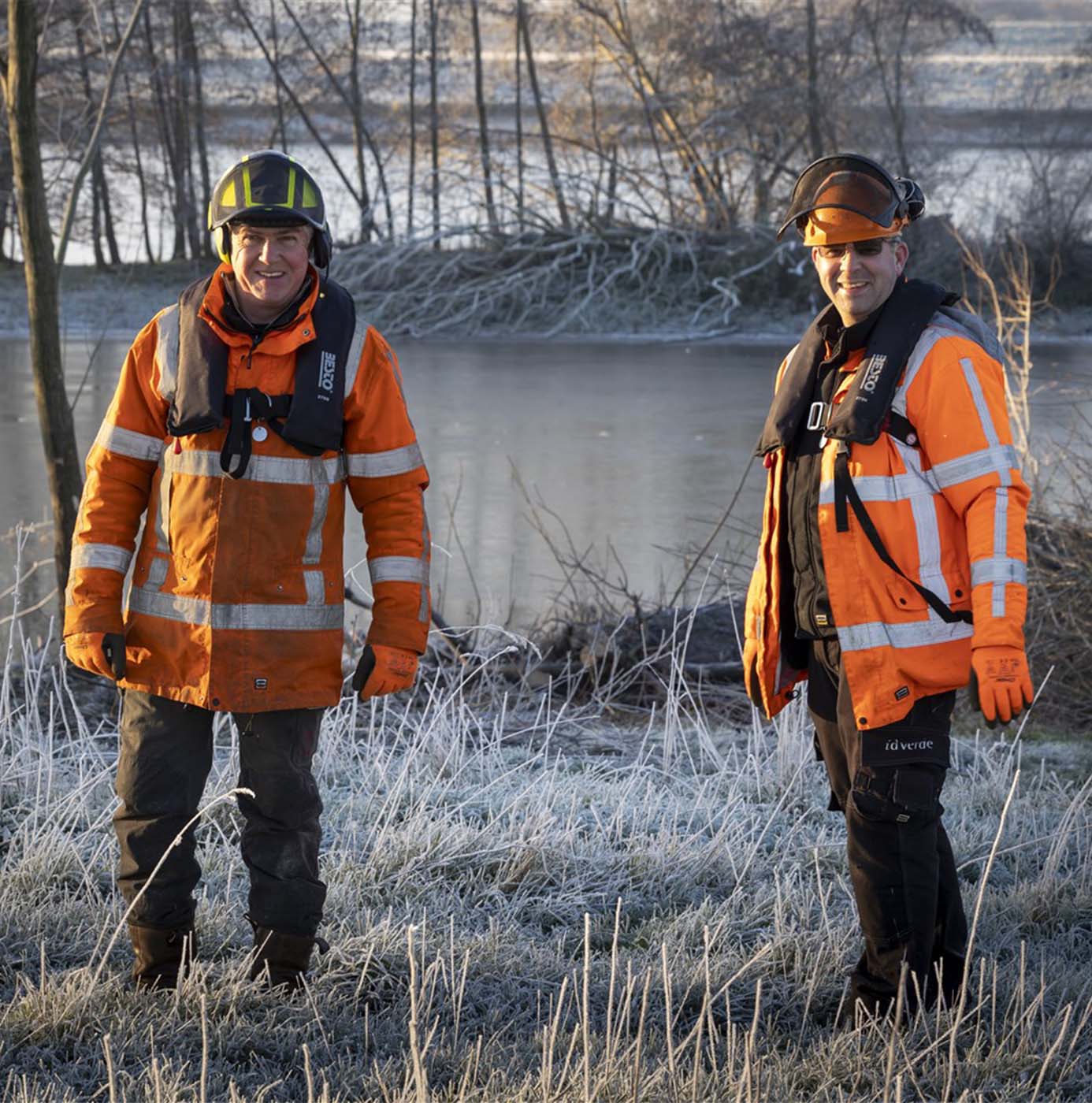 Deux agents en tenue haute visibilité et gilet de sauvetage se tiennent en bordure de rivière sur une berge givrée.