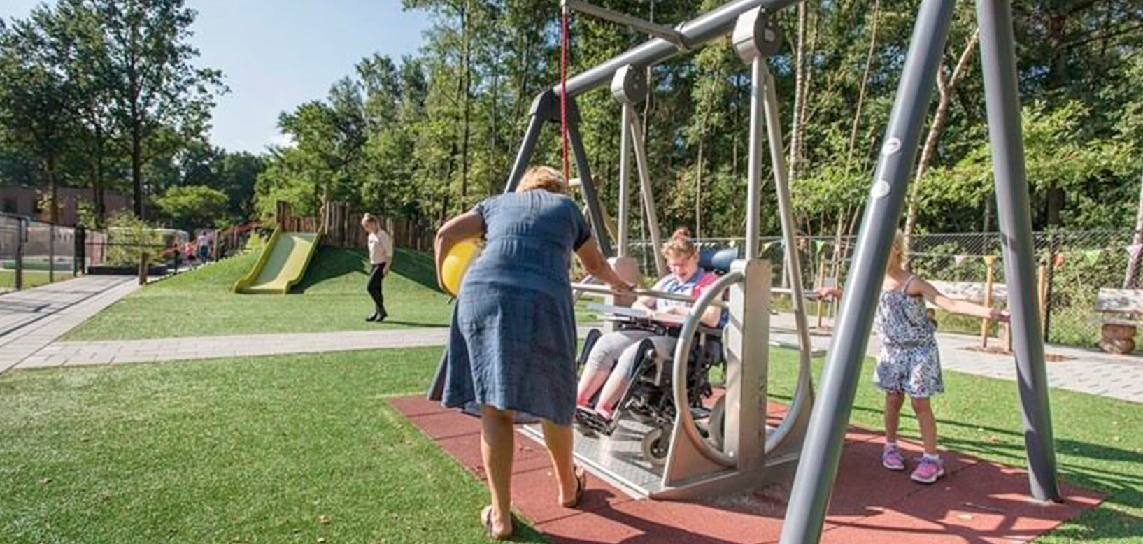 Aire de jeux inclusive avec balançoire adaptée pour fauteuil roulant, toboggan et pelouse aménagée.