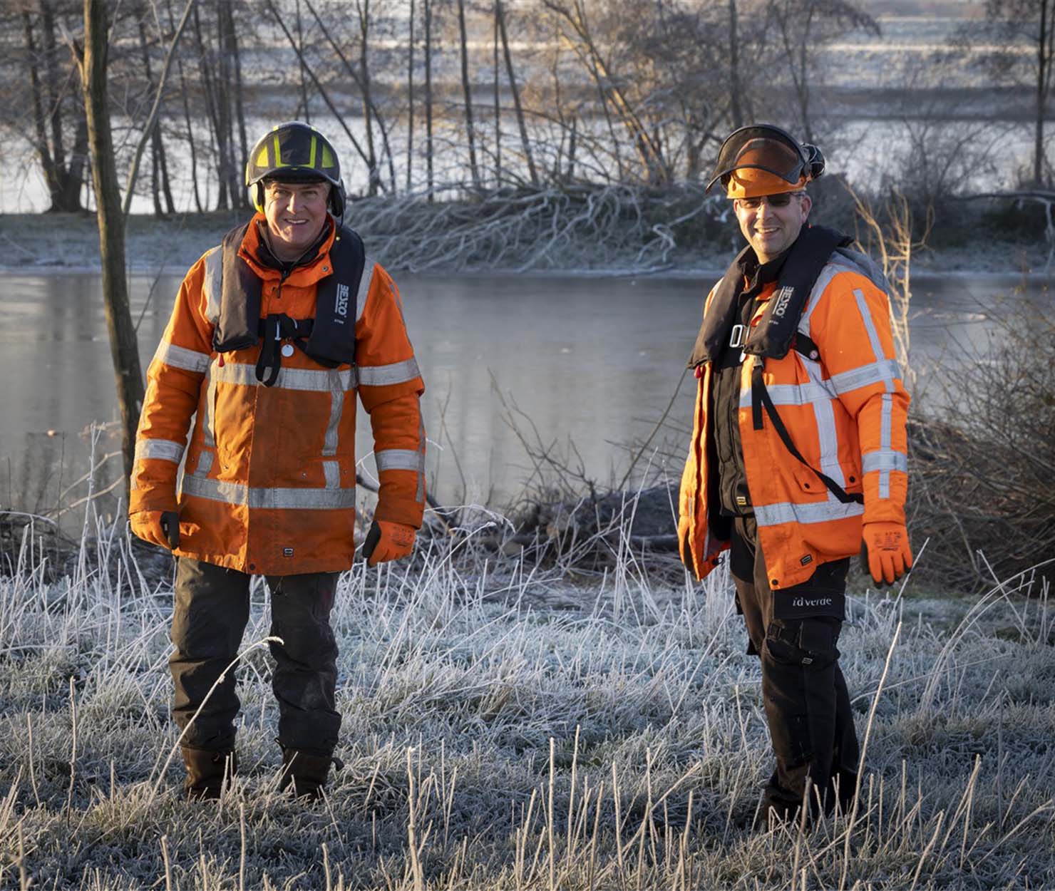 Deux agents en tenue haute visibilité et gilet de sauvetage se tiennent en bordure de rivière sur une berge givrée.