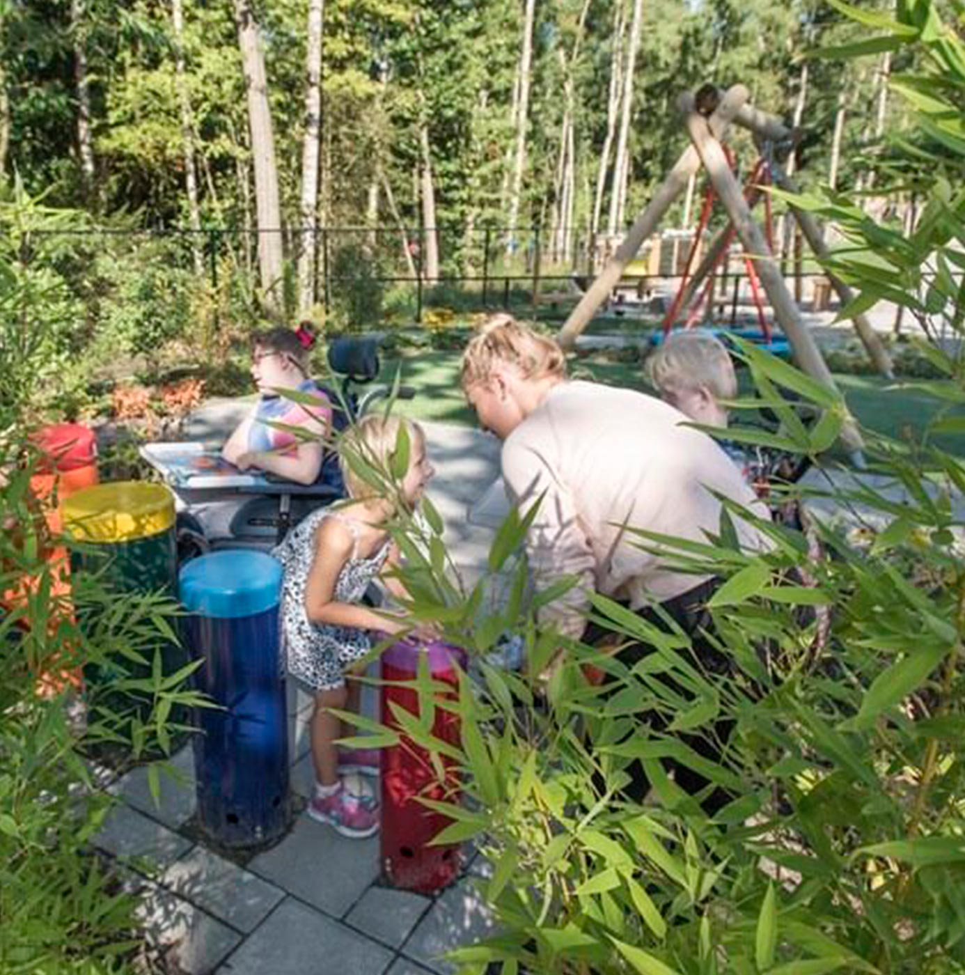 Enfants et adultes découvrent des jeux sensoriels en bambou dans un jardin arboré équipé d'une aire de jeux.