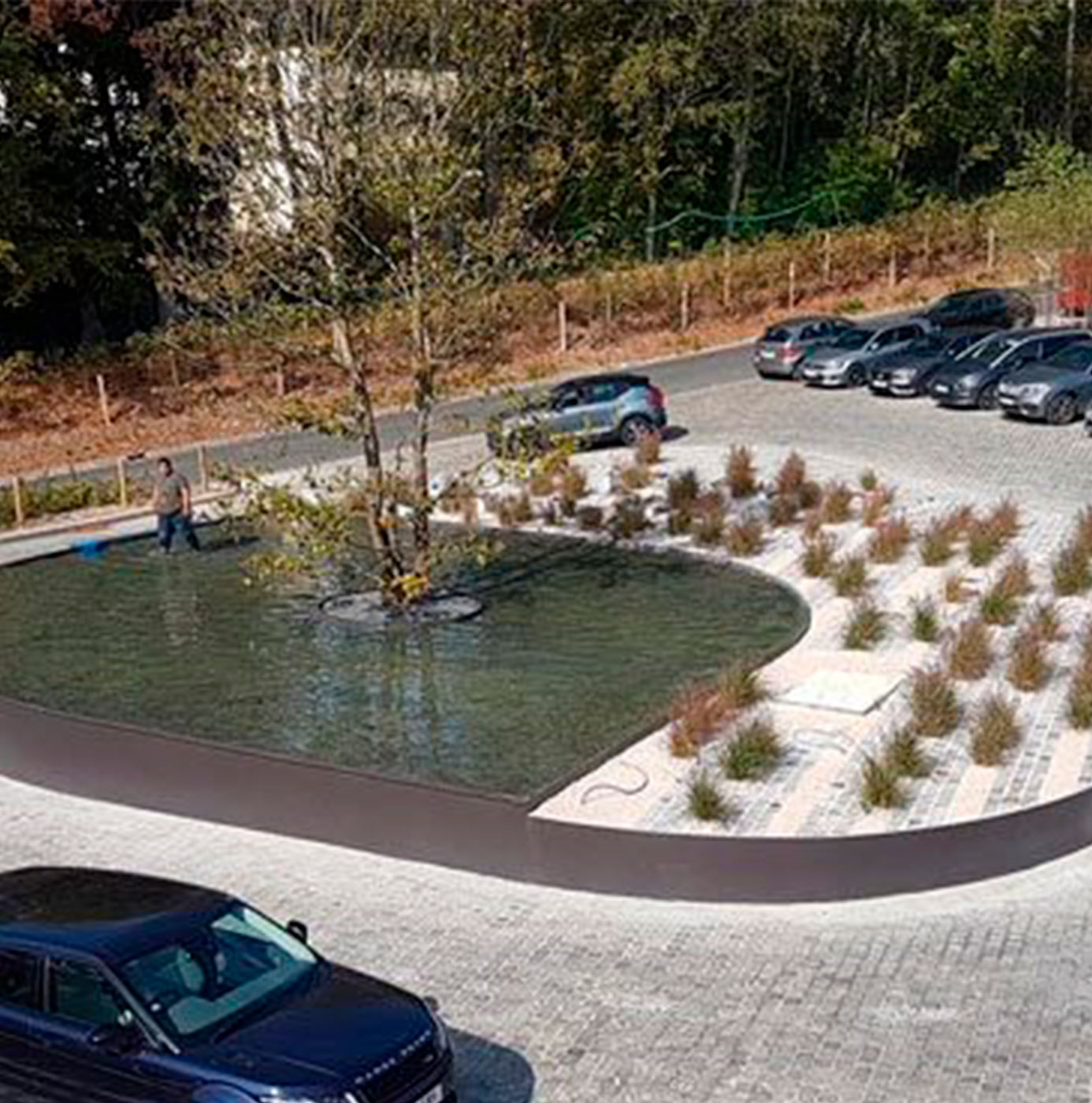 Un agent vérifie l'arrosage d'un bassin paysager planté d'un arbre au sein d'un parking.