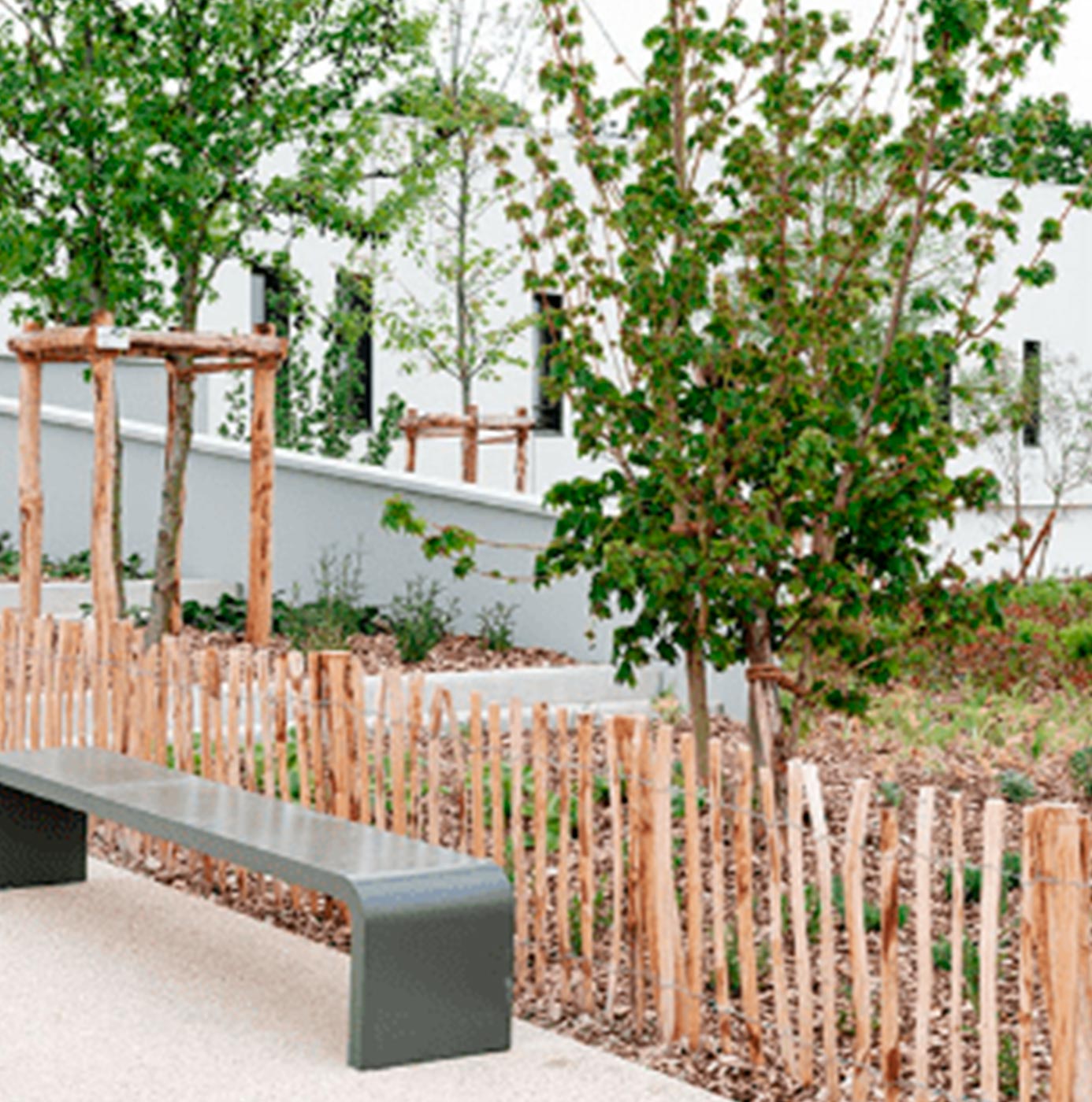 Espace vert urbain aménagé avec clôture en bois type ganivelle, arbres jeunes et banc public.