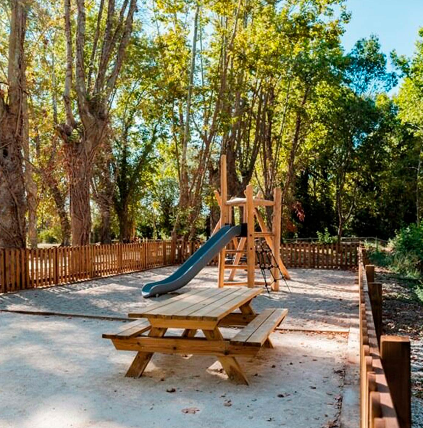 Aire de jeux extérieure clôturée avec toboggan en bois, table pique-nique et sol souple entourés d'arbres.