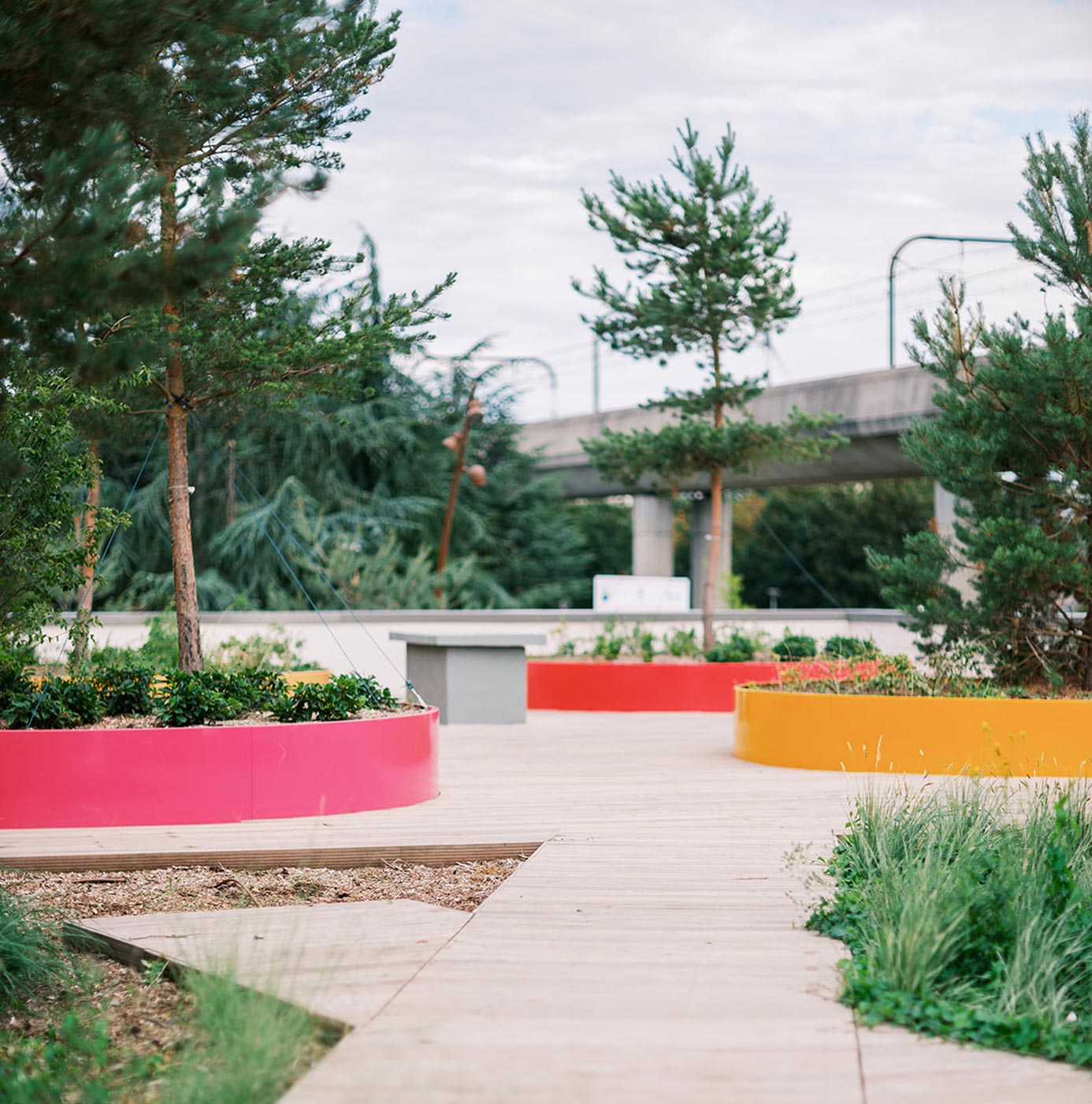 Espace paysager urbain avec bacs plantés circulaires colorés et cheminement en bois près d'un pont routier.