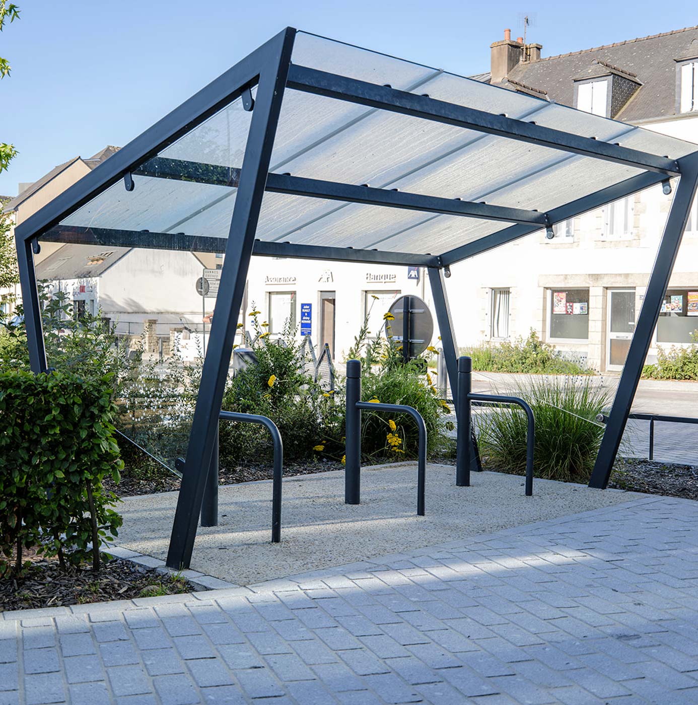 Abri à vélos aménagé sur une place, entouré de massifs de vivaces et graminées.