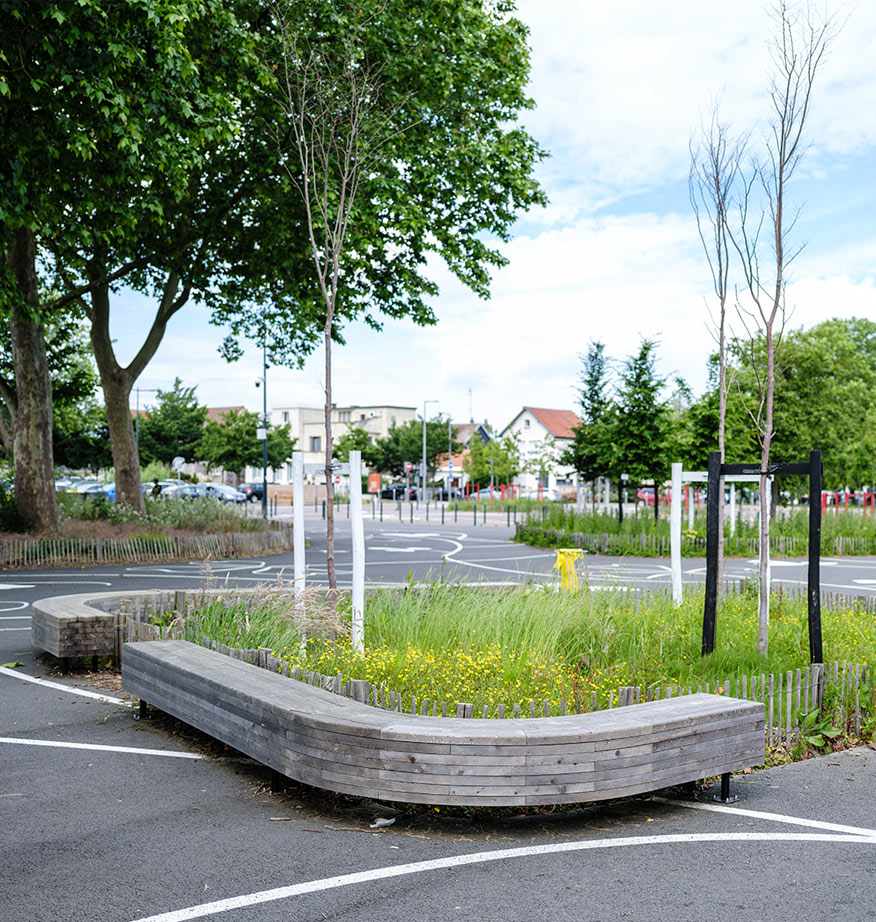 Massif fleuri et arbustif intégré dans un parking urbain, bordé d'un banc en bois incurvé.