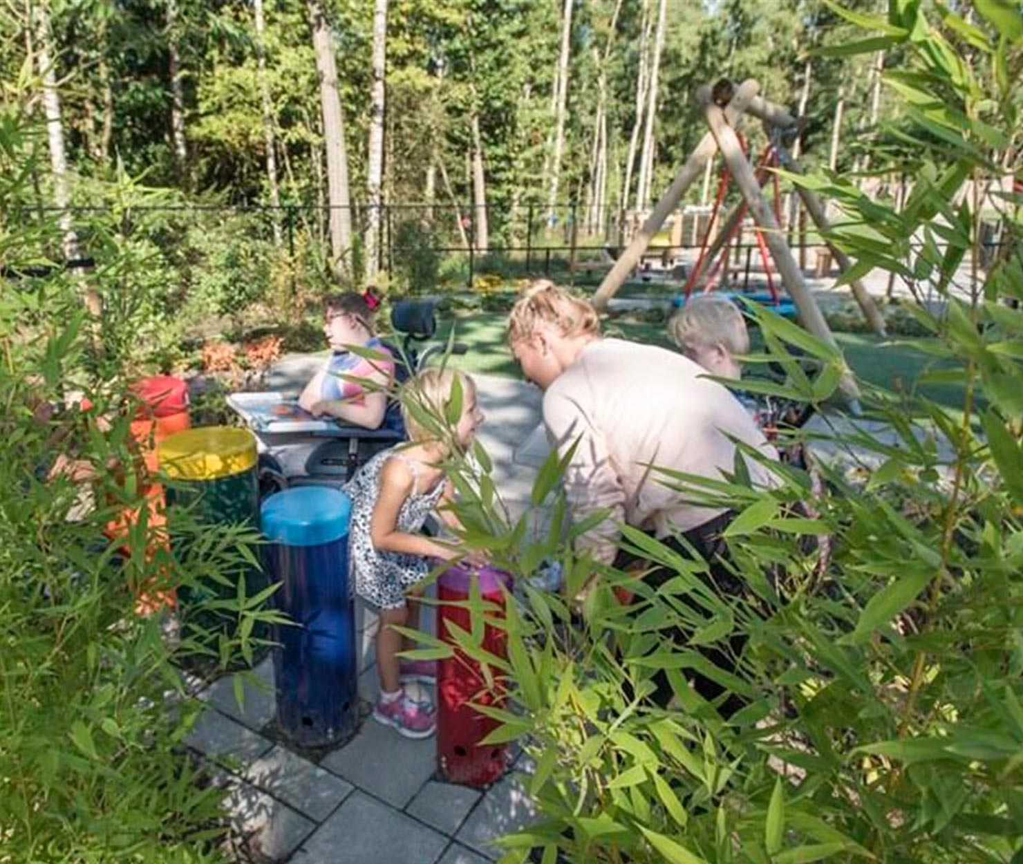 Enfants et adultes découvrent des jeux sensoriels en bambou dans un jardin arboré équipé d'une aire de jeux.