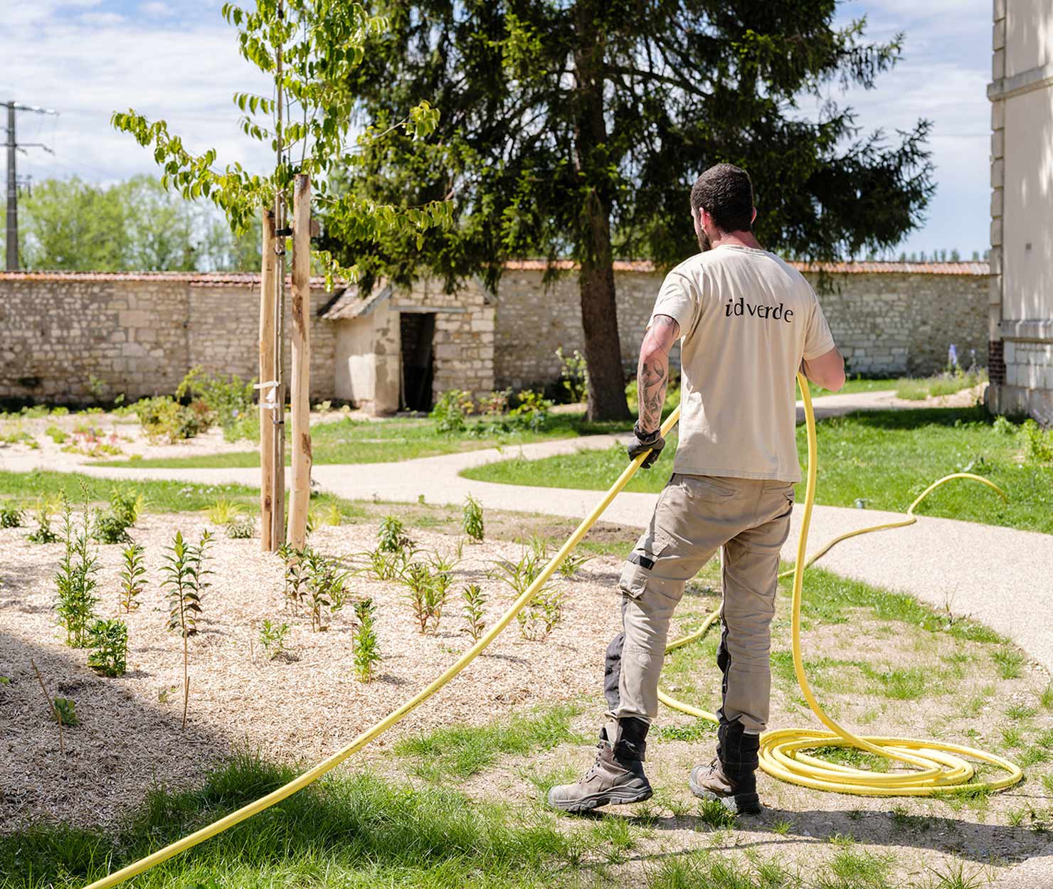 Un agent idverde déroule un tuyau d'arrosage jaune près d'un massif de jeunes plants paillés, dans un parc bordé de murs en pierre.