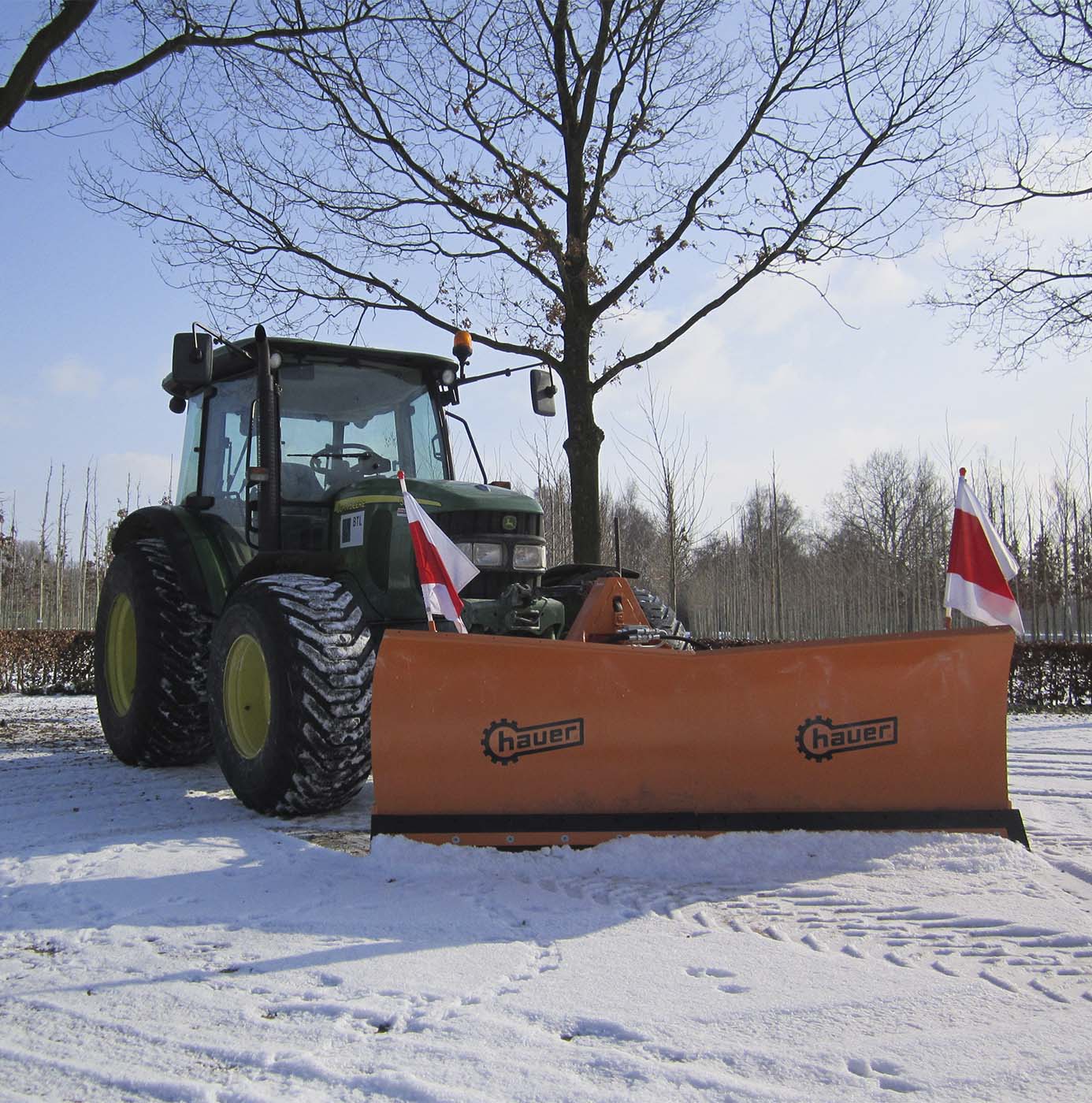 Un tracteur équipé d'une lame de déneigement dégage une allée enneigée bordée d'arbres.