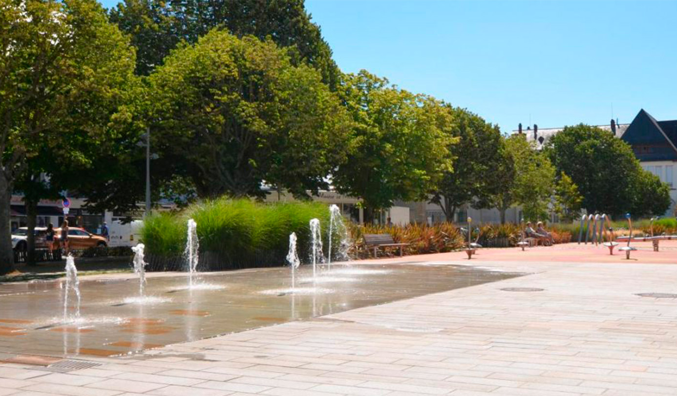 Place urbaine avec jeux d'eau, massifs de graminées et alignement d'arbres taillés.