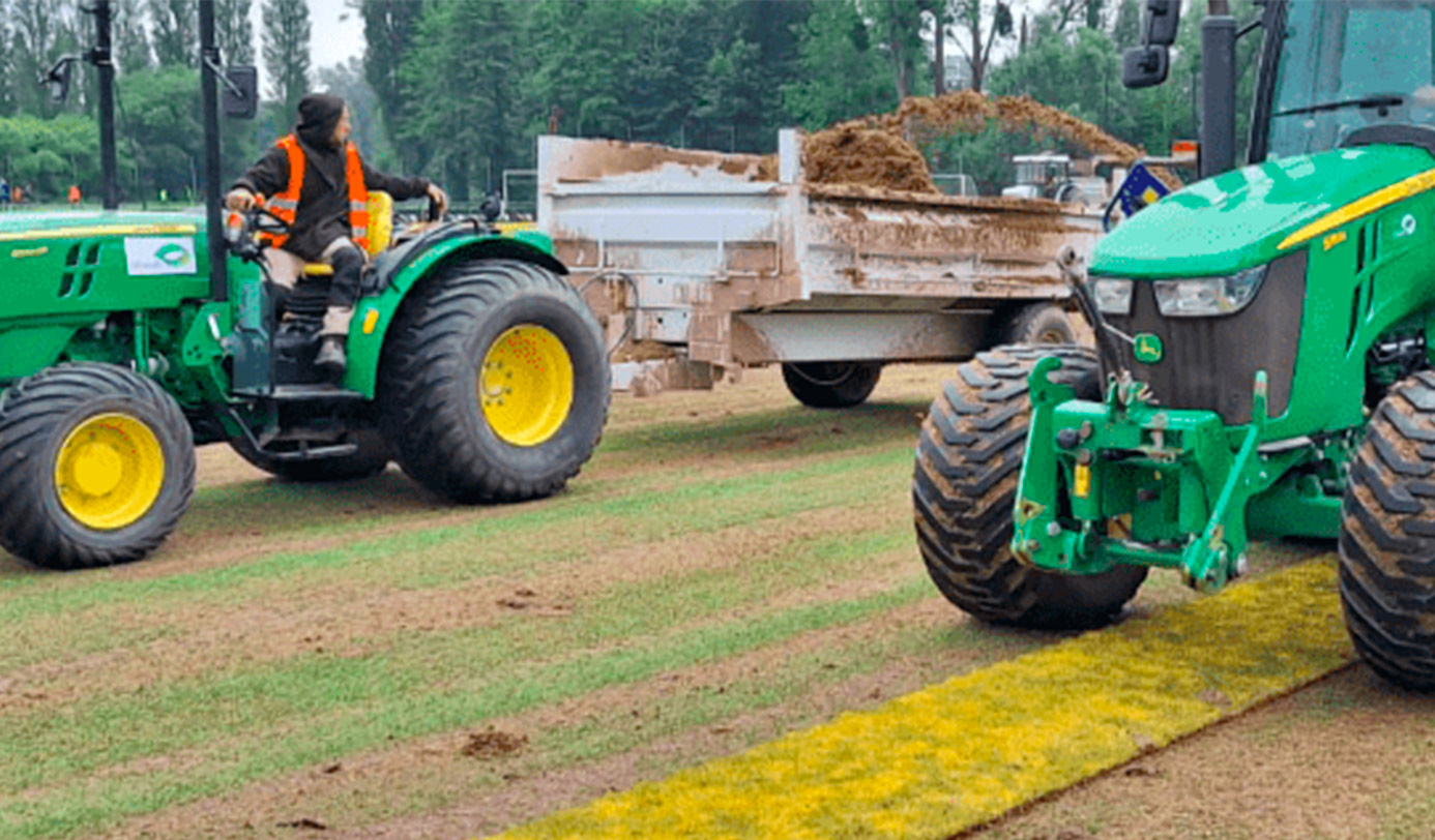 Un agent conduit un tracteur transportant du sable pour l'entretien d'un terrain de sport engazonné.