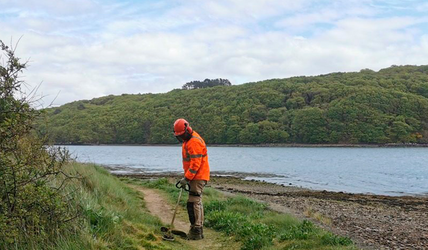 Un agent débroussaille un sentier en bord d'estuaire, équipé d'un casque et d'une tenue haute visibilité.