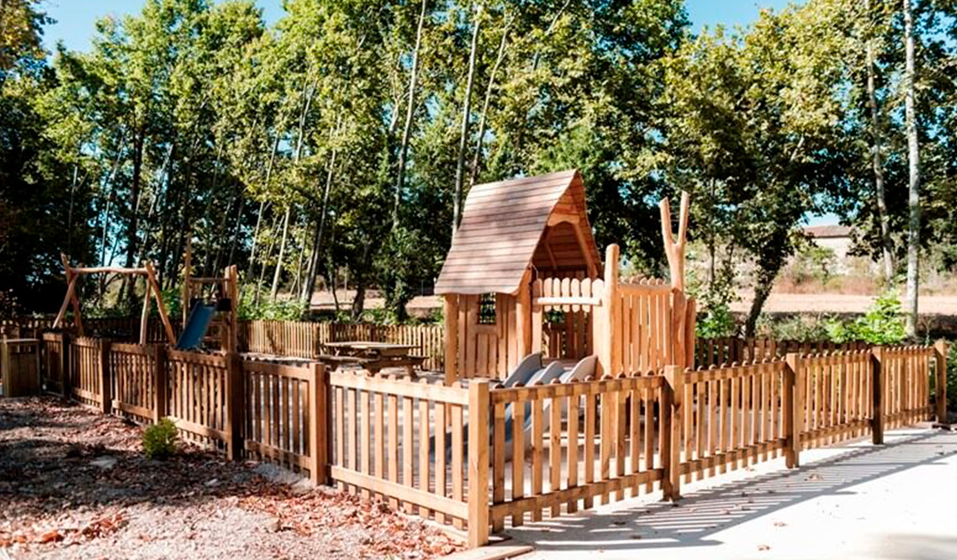 Aire de jeux en bois clôturée avec toboggan, cabane et balançoire, entourée d'arbres.
