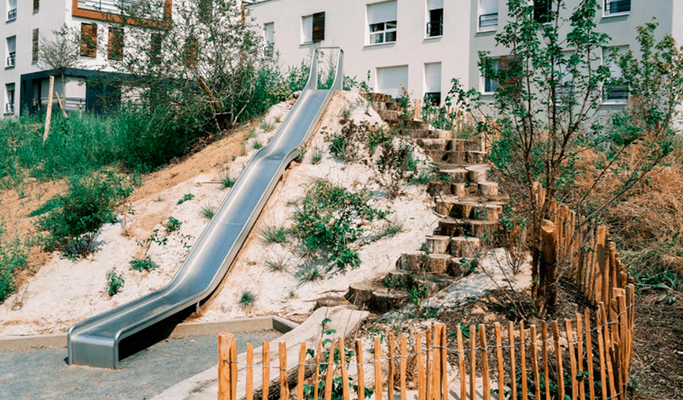 Toboggan métallique aménagé sur un talus paysager avec escalier en rondins de bois, en pied d'immeubles résidentiels.