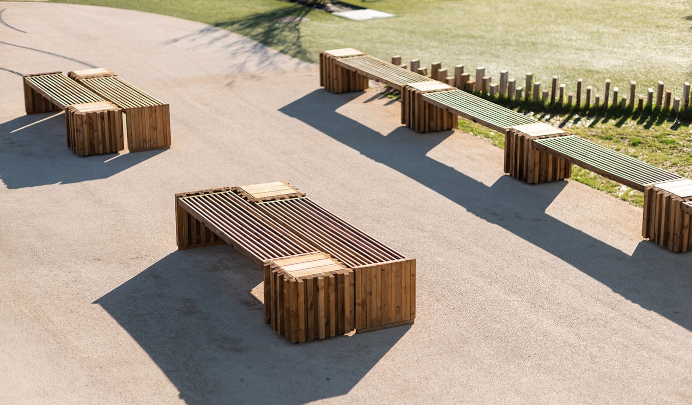 Bancs en bois design installés sur une allée minérale en bordure d'un espace vert urbain.