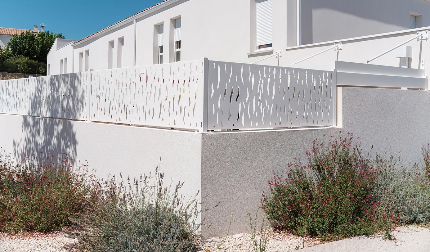 Massifs de plantes méditerranéennes plantés au pied d'un muret bordant une résidence, avec claustra en aluminium ajouré.