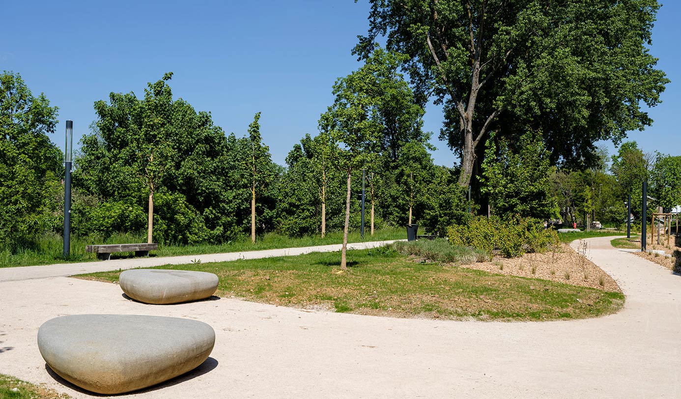 Parc paysager avec allée sablée, assises minérales en forme de galets et jeunes arbres nouvellement plantés.