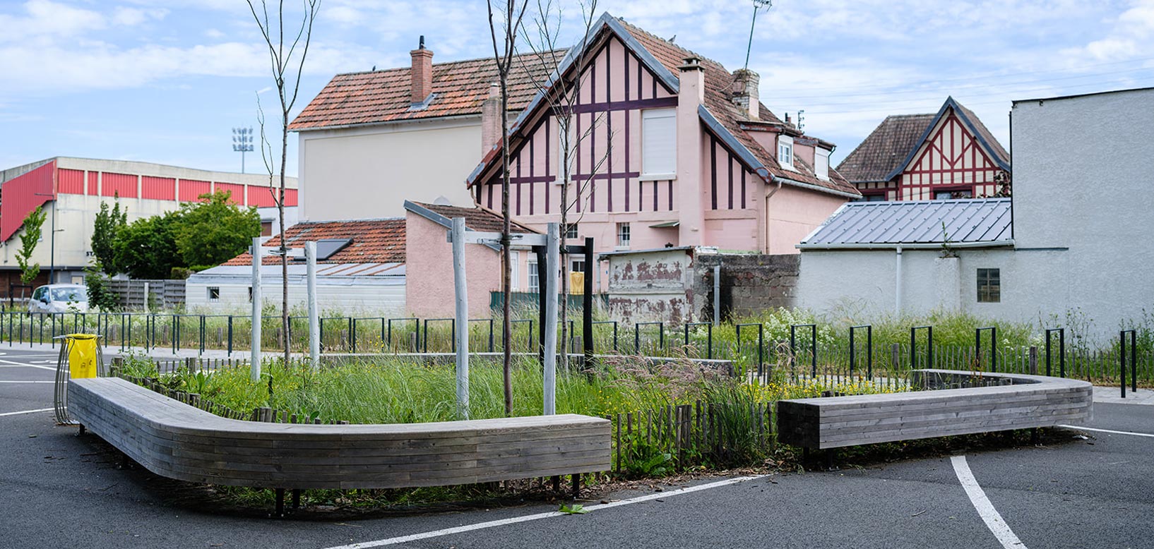 Espace paysager en milieu urbain avec massif végétalisé bordé de bancs en bois courbes, entouré de maisons à colombages.