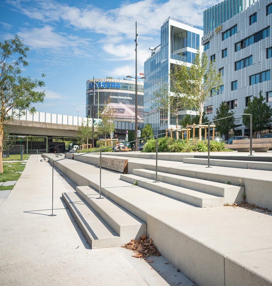 Gradins en béton et espace paysager arboré au pied d'immeubles de bureaux en milieu urbain.