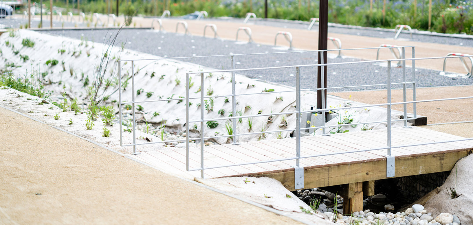 Passerelle en bois avec garde-corps enjambant une noue paysagère végétalisée en cours d'installation.