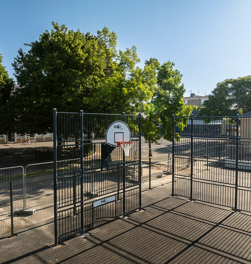 Terrain de sport clôturé avec panier de basket, entouré d'arbres dans une cour urbaine.