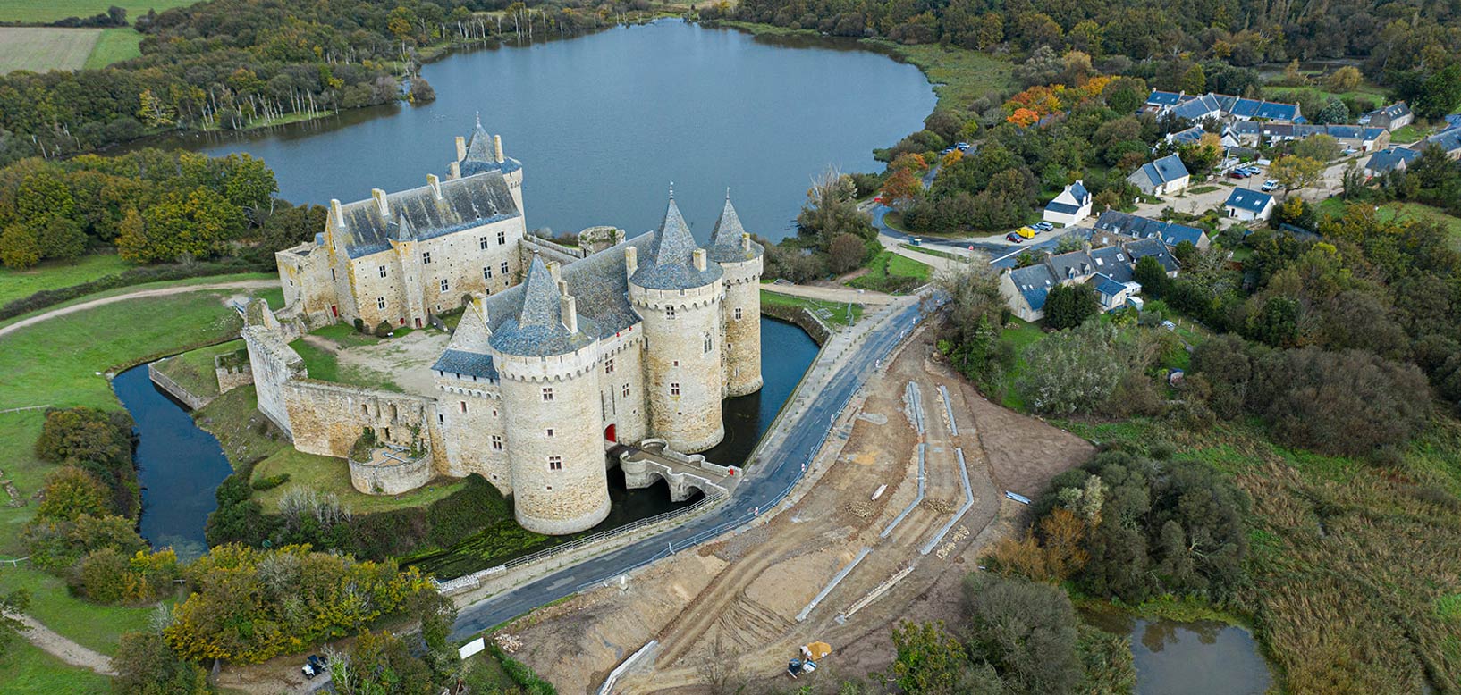 Vue aérienne d'un château entouré de douves, d'un plan d'eau et d'espaces boisés, avec des travaux d'aménagement paysager en cours à ses abords.