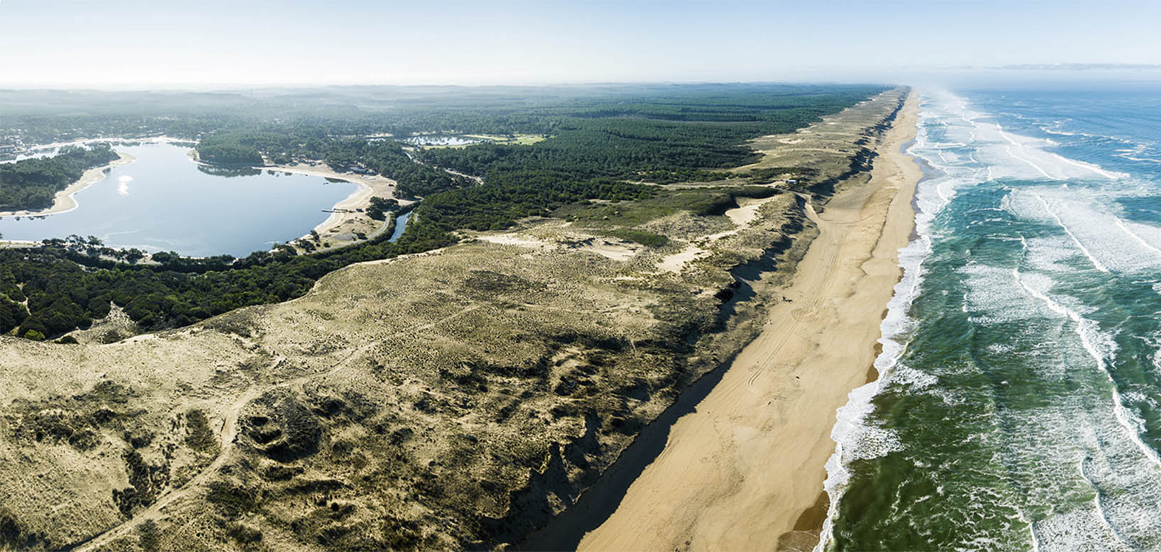 Vue aérienne d'un littoral atlantique avec dunes, forêt de pins et lac naturel en arrière-plan.