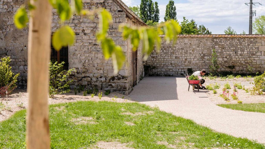 Jardinier travaillant dans un jardin historique aux murs en pierre calcaire, avec brouette rouge et plantations