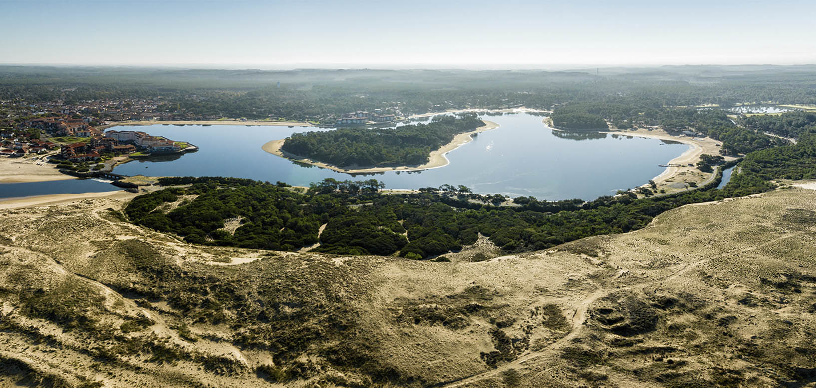 Vue aérienne d'un lac littoral bordé de dunes, de pinèdes et d'un village, sur la côte atlantique.