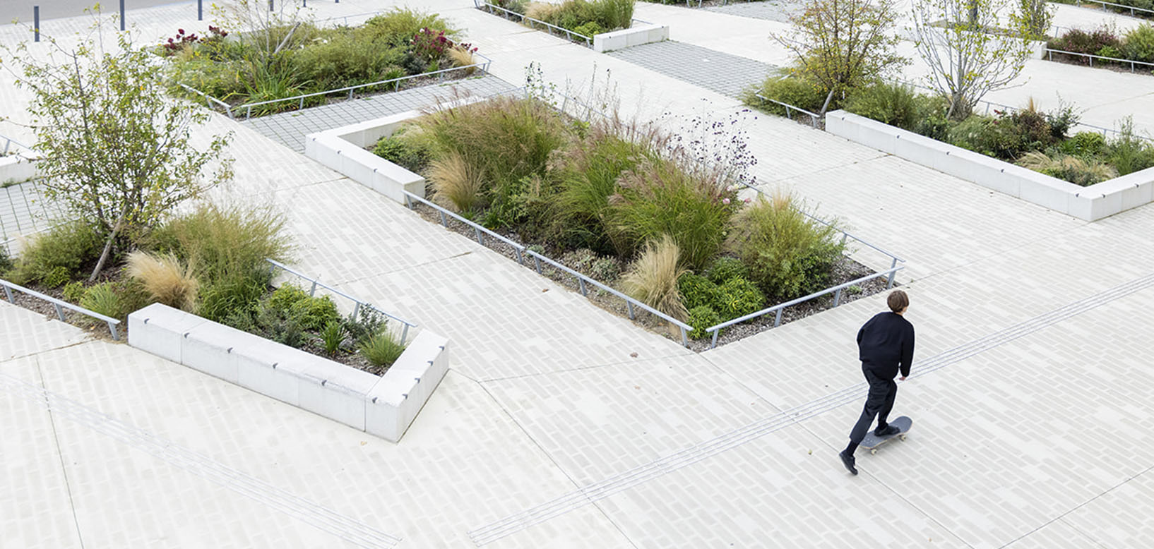 Vue aérienne d'une place minérale avec massifs de graminées et arbustes plantés en bacs, un skateur traversant l'espace.