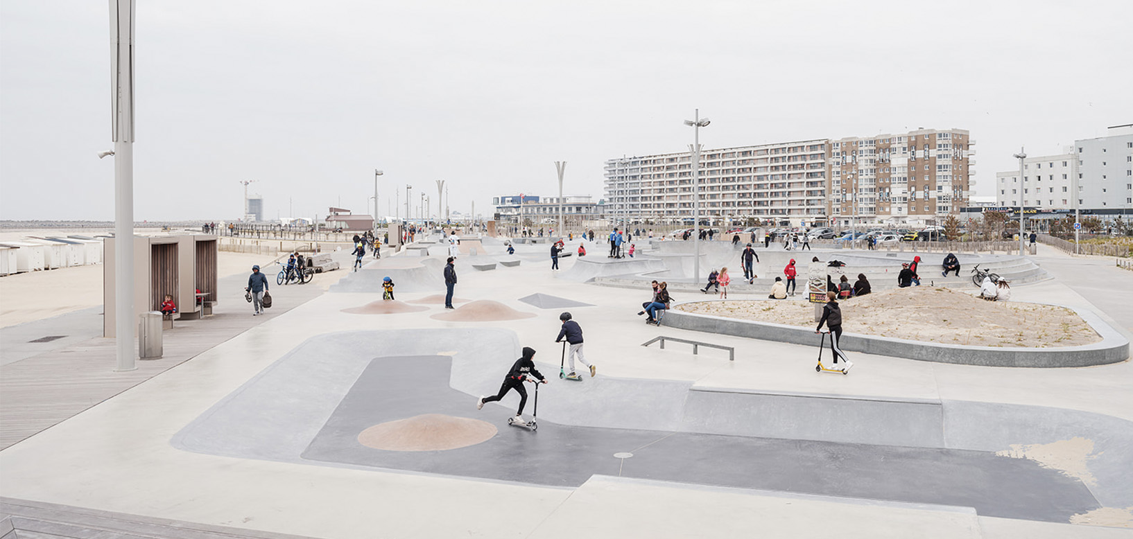 Skatepark en bord de mer avec des enfants à trottinette, en face d'un front de mer urbain.