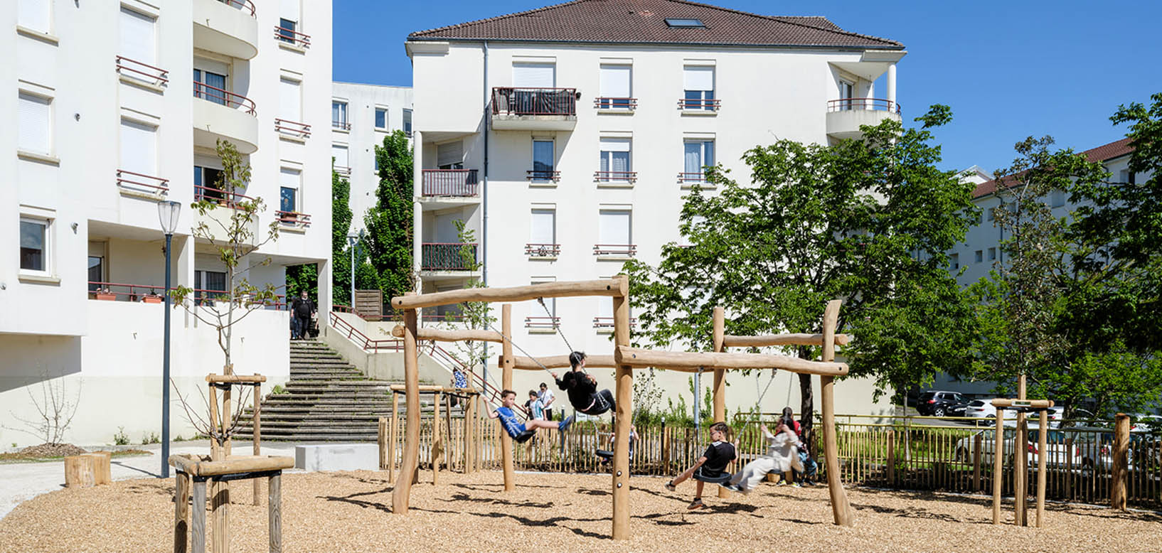 Des enfants jouent sur des balançoires en bois installées dans un espace vert au pied d'immeubles résidentiels.