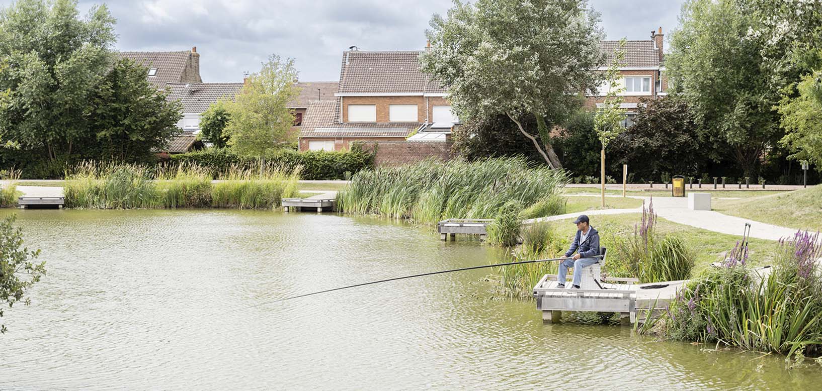 Un pêcheur assis sur un ponton en bois au bord d'un étang aménagé en milieu urbain, entouré de roselières.