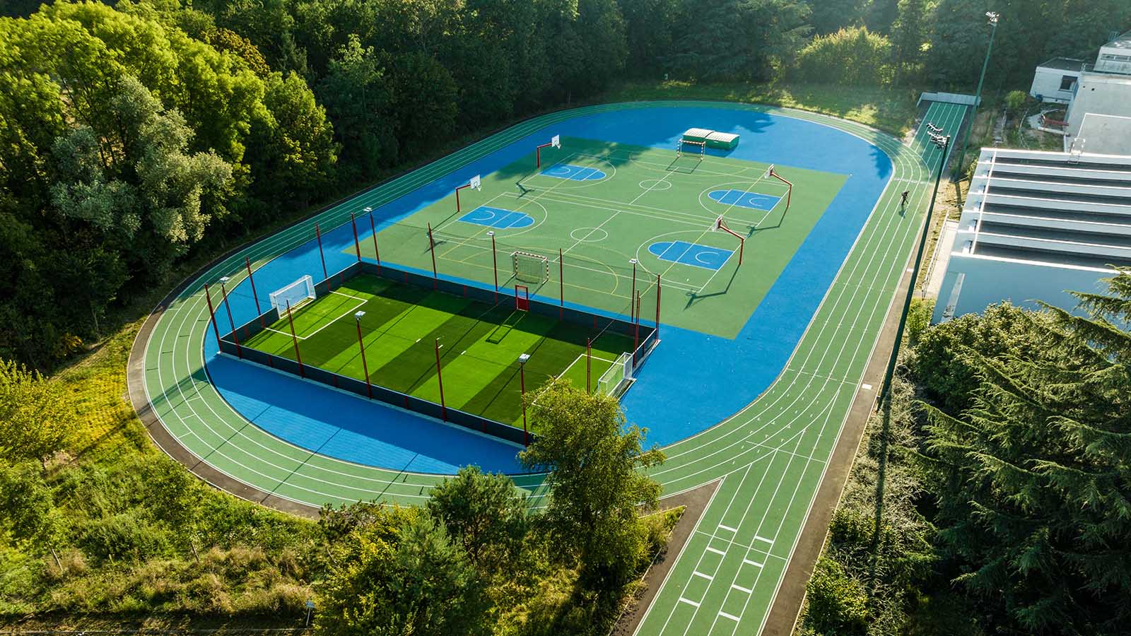 Vue aérienne d'un complexe sportif moderne avec piste d'athlétisme bleue, terrain de football synthétique et courts multisports