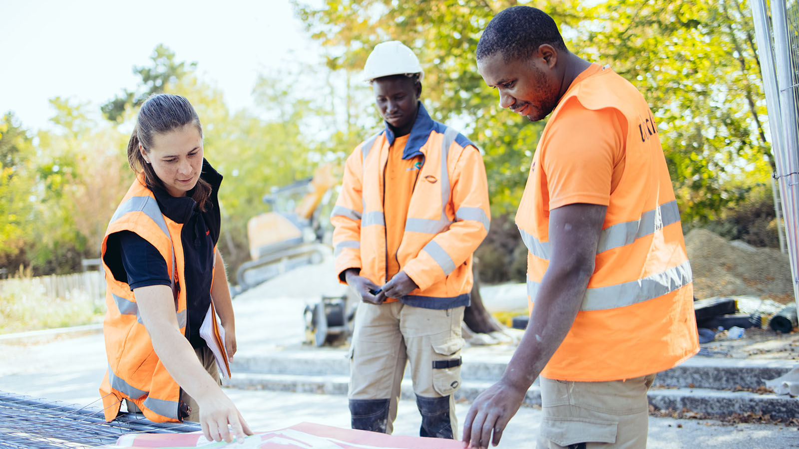 Trois agents en gilets orange fluorescent examinent des plans sur un chantier de travaux extérieurs