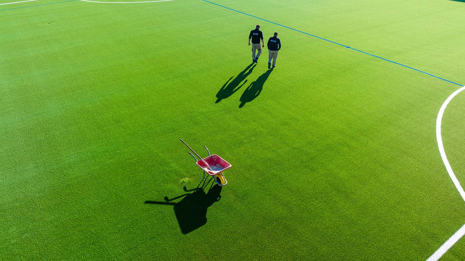 Vue aérienne de deux techniciens idverde inspectant un terrain de sport en gazon synthétique vert avec une brouette rouge