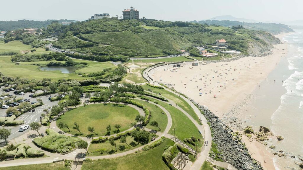 Vue aérienne d'un parc urbain aménagé en bord de mer avec une plage fréquentée et une colline verdoyante