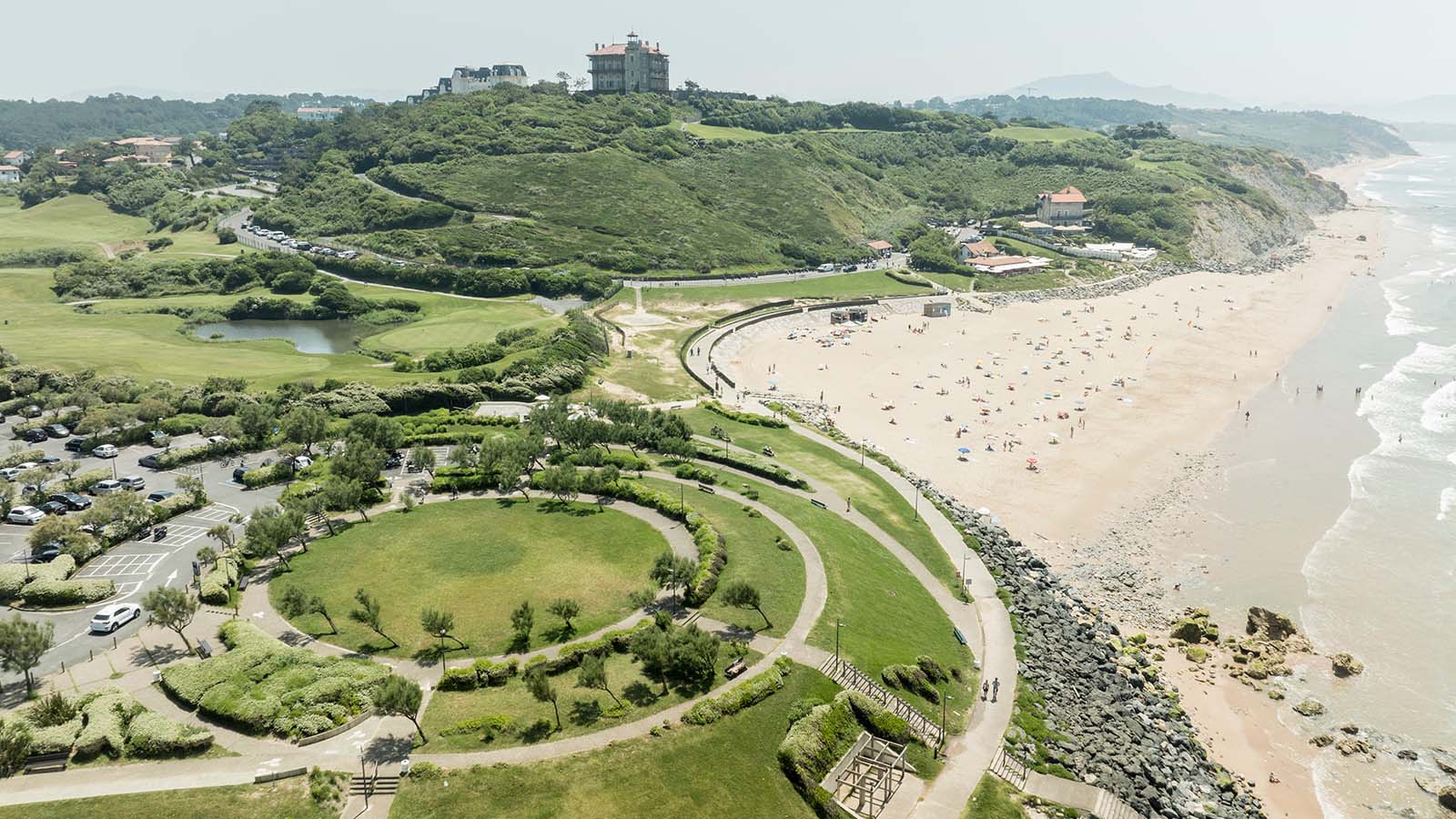 Vue aérienne d'un parc urbain aménagé en bord de mer avec une plage fréquentée et une colline verdoyante